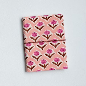 May include: A pink floral patterned journal with a brown string closure. The cover features a repeating pattern of pink flowers with brown stems and leaves on a light pink background. The journal is rectangular and appears to be closed.