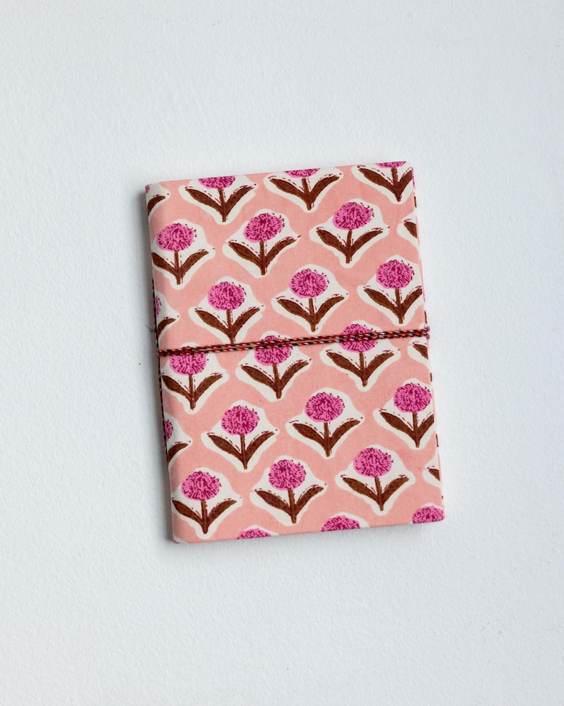 May include: A pink floral patterned journal with a brown string closure. The cover features a repeating pattern of pink flowers with brown stems and leaves on a light pink background. The journal is rectangular and appears to be closed.