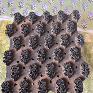 May include: A wooden block with a floral design, featuring rows of stylized flowers. The block is a dark brown color, and the flowers are carved in a detailed pattern. The background shows a fabric with a repeating floral pattern in shades of green and purple.