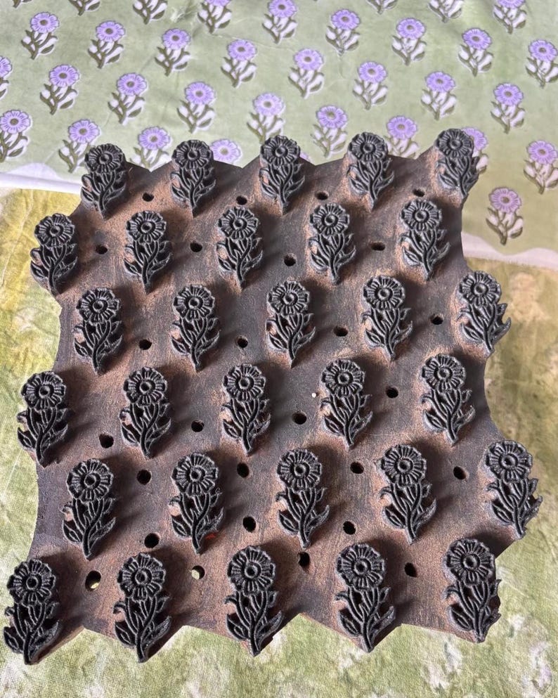 May include: A wooden block with a floral design, featuring rows of stylized flowers. The block is a dark brown color, and the flowers are carved in a detailed pattern. The background shows a fabric with a repeating floral pattern in shades of green and purple.