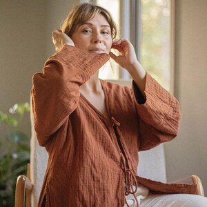 May include: A rust-colored, long-sleeved wrap top with a textured, crinkled fabric. The top features a V-neck and tie closure. The model is wearing the top with white pants while seated in a wooden chair.