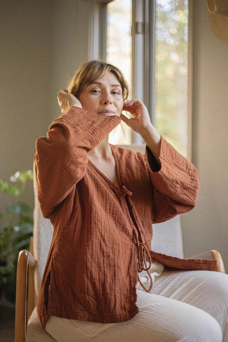 May include: A rust-colored, long-sleeved wrap top with a textured, crinkled fabric. The top features a V-neck and tie closure. The model is wearing the top with white pants while seated in a wooden chair.