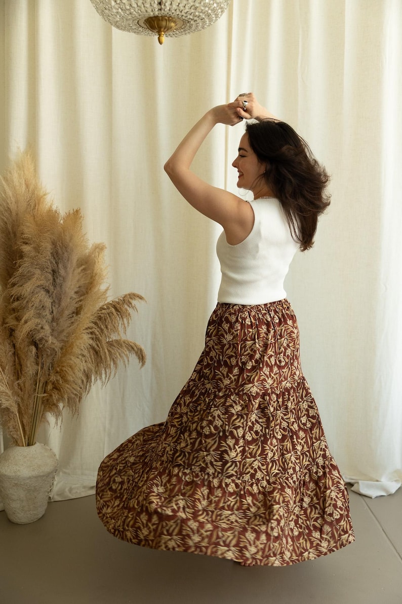 May include: A woman wearing a white ribbed tank top and a long, flowing skirt. The skirt features a repeating floral pattern in shades of brown and cream. The woman is dancing with her arms raised.