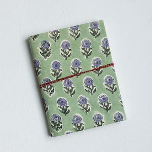 May include: A rectangular notebook wrapped in green paper with a repeating pattern of purple flowers. The notebook is secured with a red and white twine. The background is plain white.