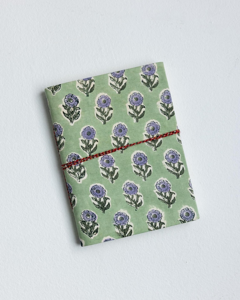 May include: A rectangular notebook wrapped in green paper with a repeating pattern of purple flowers. The notebook is secured with a red and white twine. The background is plain white.