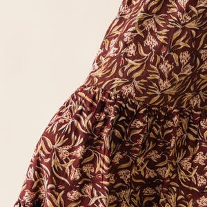 May include: Close-up of a tiered skirt with a burgundy background and a repeating floral pattern in cream and gold. The fabric has a soft, flowing appearance, suggesting a comfortable garment.