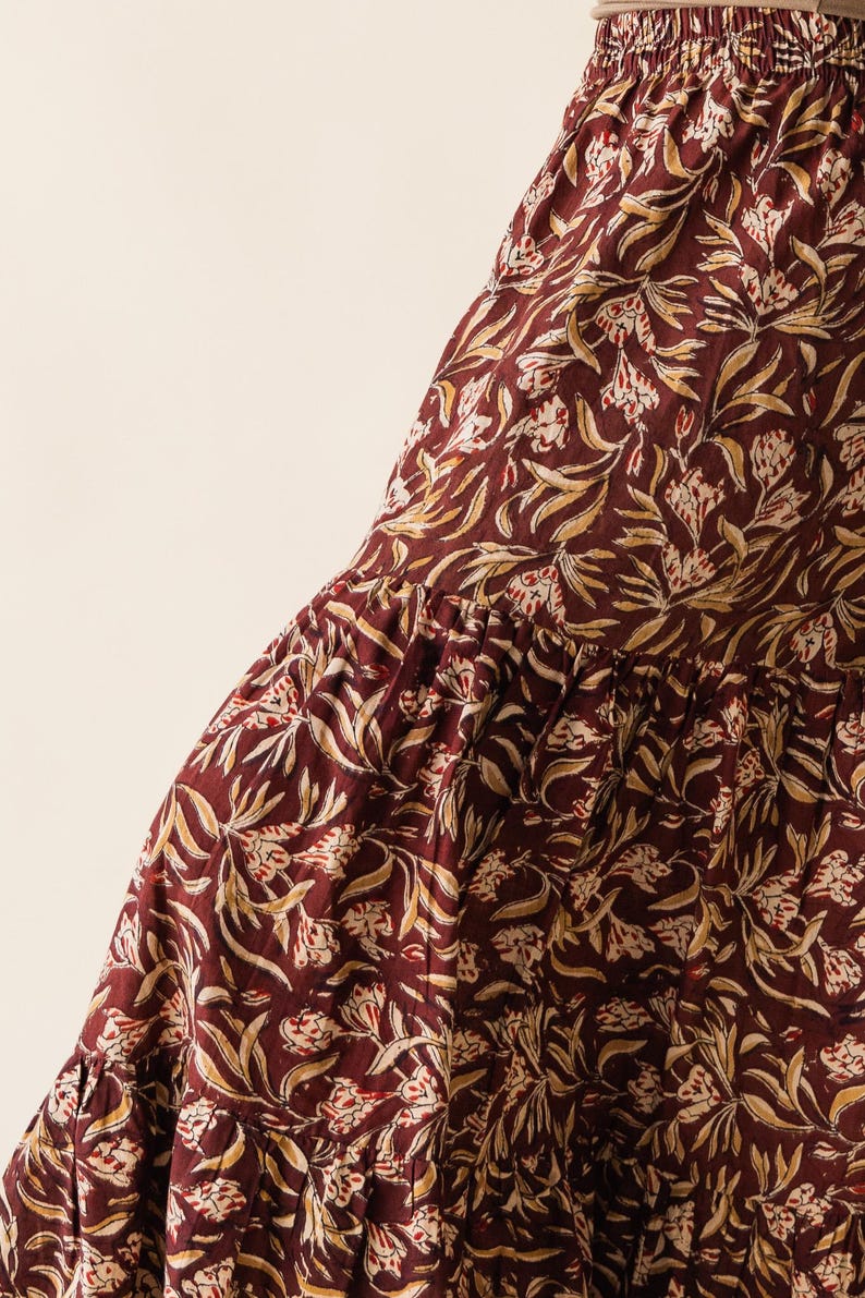 May include: Close-up of a tiered skirt with a burgundy background and a repeating floral pattern in cream and gold. The fabric has a soft, flowing appearance, suggesting a comfortable garment.
