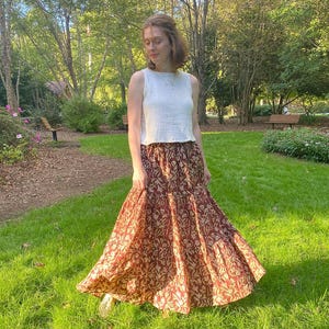 May include: A woman wearing a white sleeveless top and a long, flowing skirt. The skirt features a maroon background with a repeating floral pattern in a lighter tan color. The outfit is worn outdoors in a park setting.