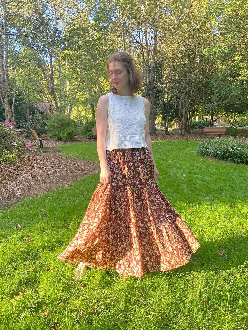 May include: A woman wearing a white sleeveless top and a long, flowing skirt. The skirt features a maroon background with a repeating floral pattern in a lighter tan color. The outfit is worn outdoors in a park setting.