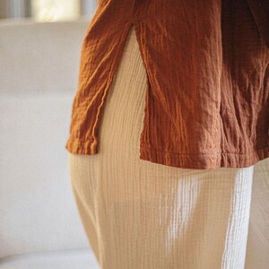 May include: A close-up of a rust-colored top with a side slit, paired with cream-colored pants. The fabric appears to be textured and lightweight, suggesting a casual and comfortable style. The outfit is simple and elegant.
