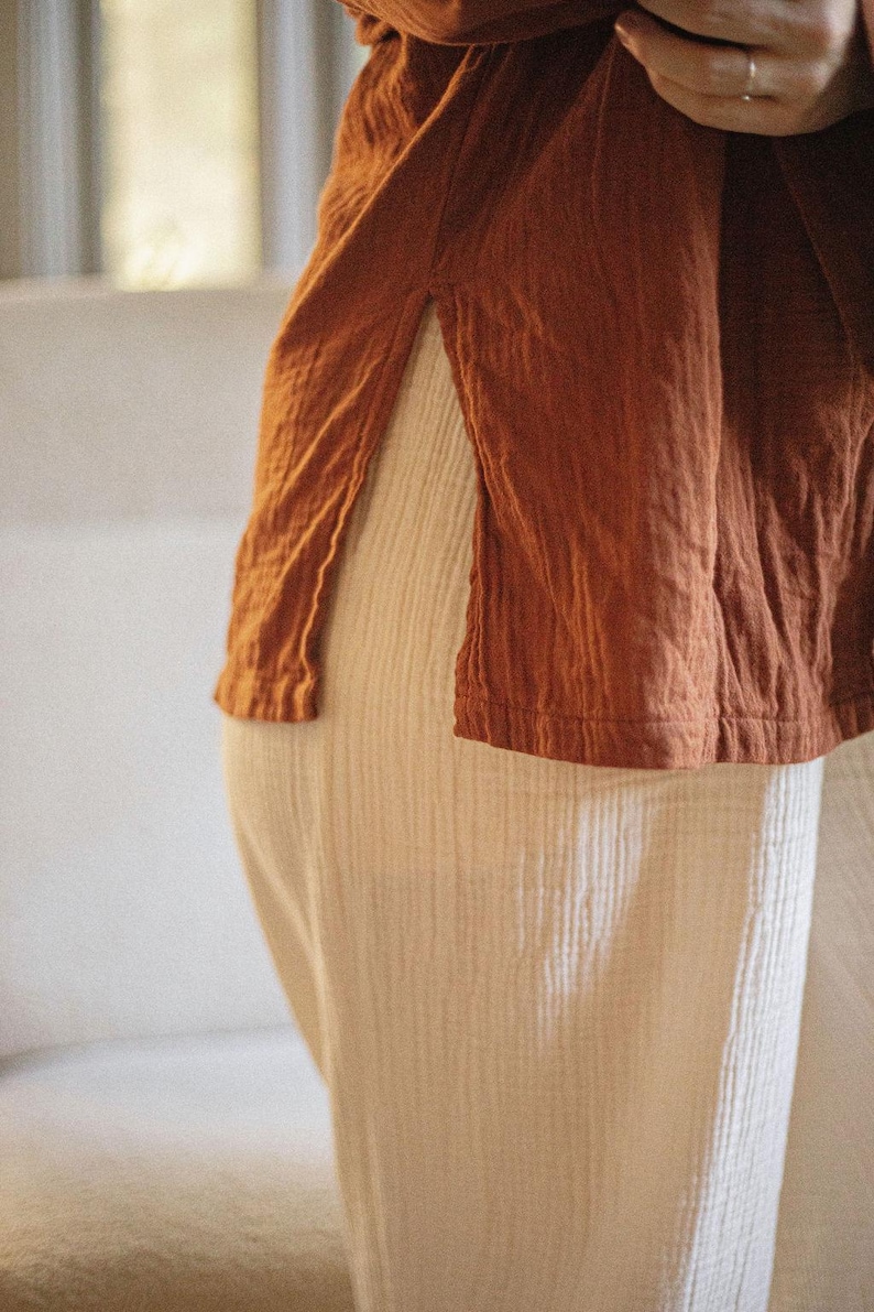 May include: A close-up of a rust-colored top with a side slit, paired with cream-colored pants. The fabric appears to be textured and lightweight, suggesting a casual and comfortable style. The outfit is simple and elegant.