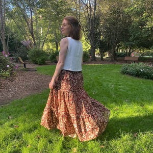 May include: A sleeveless white top paired with a long, flowing skirt. The skirt features a tiered design with a pattern of brown and beige floral elements. The outfit is worn outdoors in a park setting with green grass and trees.