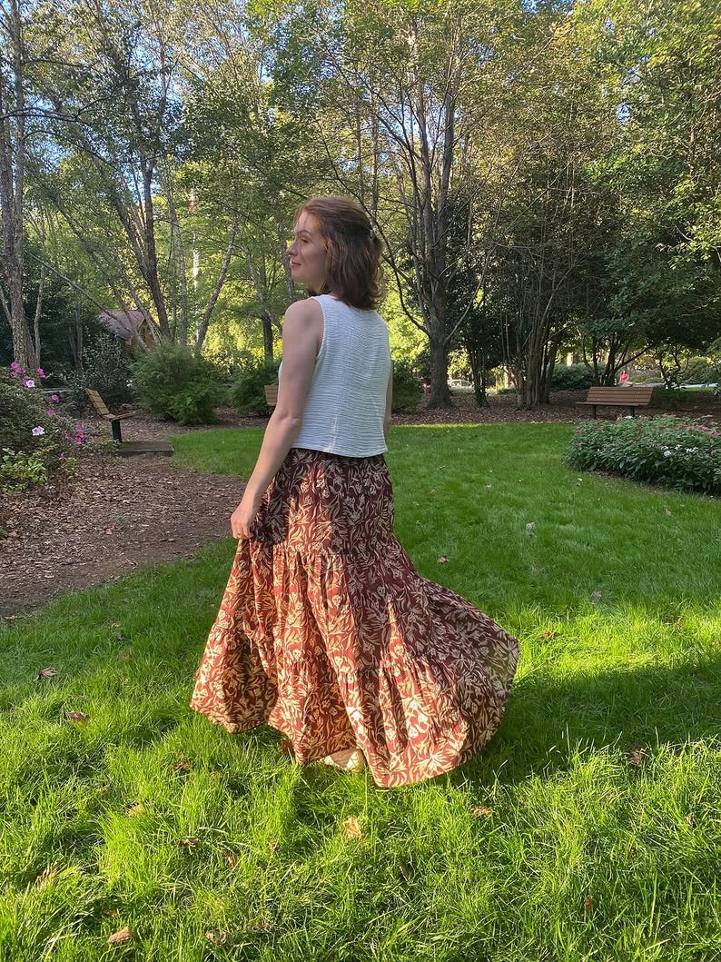 May include: A sleeveless white top paired with a long, flowing skirt. The skirt features a tiered design with a pattern of brown and beige floral elements. The outfit is worn outdoors in a park setting with green grass and trees.