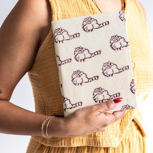 May include: A cream-colored notebook featuring a repeating pattern of cartoon raccoons in dark brown ink. The notebook is held by a person wearing a yellow outfit. The raccoons have glasses and long tails.