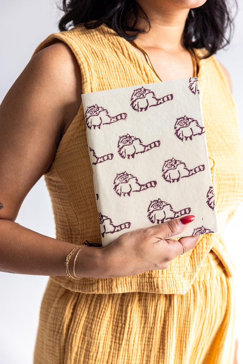 May include: A cream-colored notebook featuring a repeating pattern of cartoon raccoons in dark brown ink. The notebook is held by a person wearing a yellow outfit. The raccoons have glasses and long tails.