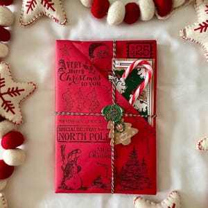 Könnte beinhalten: Ein rotes Geschenk, das in festliches Papier mit einem Weihnachtsmotiv verpackt ist. Das Papier zeigt einen Schneemann, einen Weihnachtsbaum und den Text "A VERY Merry Christmas to you". Das Geschenk ist mit Schnur und einem grünen Wachssiegel gebunden. Ein kleines Etikett mit dem Text "Merry Christmas" ist an der Schnur befestigt.