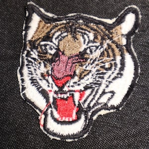 May include: A close-up of an embroidered tiger head patch. The tiger has a white face with black stripes and a red open mouth. The tiger's eyes are black and the nose is pink.