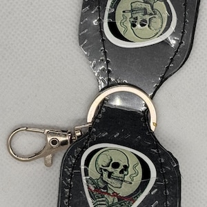 May include: A black leather keychain with a silver clasp and ring. Each piece features a skull graphic with a cigarette. The keychain is held together with silver rivets and stitching.