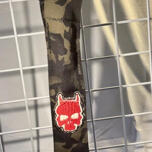 May include: A camouflage drumstick holder with a red skull patch. The holder is made of durable fabric and features a black strap for securing the drumsticks. The drumsticks are made of light-colored wood.