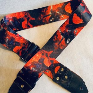 May include: A black guitar strap with a red and black print of a band member playing a guitar. The strap has black leather ends with metal buckles.
