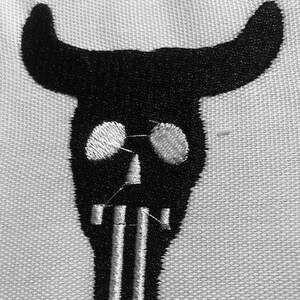 May include: Black and white embroidered bull skull design. The skull has white eyes and a white mouth with three vertical lines.