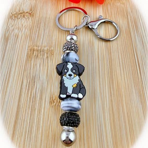 May include: A keychain featuring a central silicone charm of a black, white, and tan dog. The charm is flanked by alternating beads, including black rhinestone-encrusted spheres, smooth grey and white marbled beads, and polished silver-tone metal beads.