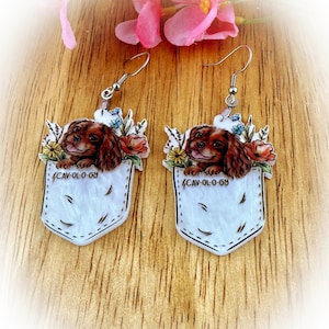 May include: A pair of dangle earrings featuring a Cavalier King Charles Spaniel dog design. Each earring has a white pocket shape with a dog's head peeking out, surrounded by flowers. The text "CAV-OL-0-68" is printed on each earring.