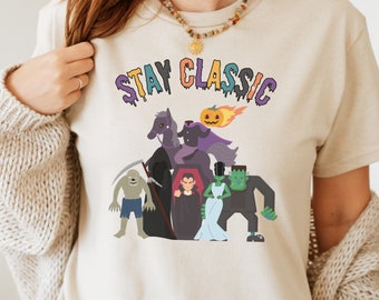 May include: A beige short-sleeve t-shirt featuring the text STAY CLASSIC in colorful, dripping letters above a graphic illustration of various classic horror movie monsters, including a headless horseman, a vampire, and a green creature.