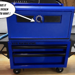 May include: A blue metal tool chest with black drawers and side trays. The front features a gear-shaped emblem with a wrench icon and the text CUSTOMIZE IT WITH ANY DESIGN OR WORD YOU WANT. The unit includes black wheels and U.S. GENERAL branding.