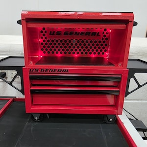 May include: A red U.S. GENERAL rolling tool chest with black accents. The chest has multiple drawers, a top compartment, and side trays. The U.S. GENERAL logo is visible on the front. The interior is illuminated with red lights.