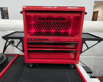 May include: A red U.S. GENERAL rolling tool chest with black accents. The chest has multiple drawers, a top compartment, and side trays. The U.S. GENERAL logo is visible on the front. The interior is illuminated with red lights.
