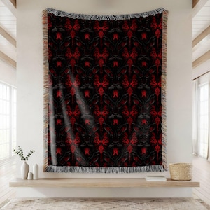 May include: A black and red patterned throw blanket with a fringed edge. The design features a repeating floral motif in shades of red against a black background. The blanket is displayed hanging on a wall.