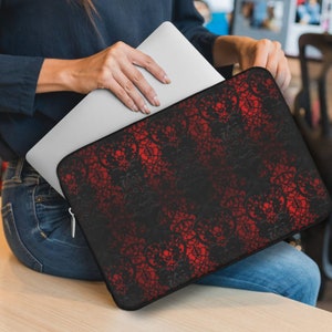 May include: A black and red laptop sleeve with a decorative pattern. The sleeve is held by a person wearing a dark blue shirt and jeans. The pattern is a repeating design of swirling shapes. The laptop is silver.