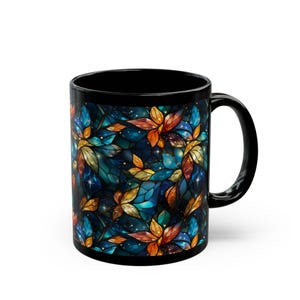 May include: Black ceramic mug featuring a stained-glass floral design in shades of blue, orange, and yellow. The design includes a starry background. The mug has a black handle and interior.