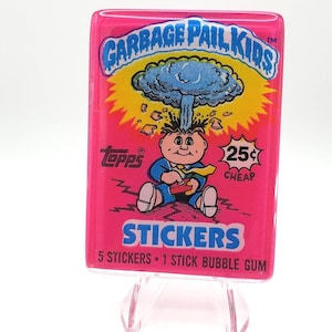 Könnte beinhalten: Eine rechteckige Garbage Pail Kids Stickerpackung mit leuchtend rosa Hintergrund. Die Grafik zeigt eine Cartoon-Figur mit einer aus dem Kopf aufsteigenden Atompilzwolke, die einen blauen Anzug trägt. Der Text umfasst Topps, 25 cents und 5 stickers 1 stick bubble gum.