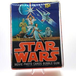 May include: A rectangular Star Wars movie photo cards bubble gum box featuring a 1977 illustration of Luke Skywalker, Princess Leia, C-3PO, R2-D2, an X-wing fighter, and the Death Star against a blue space background with the text 15 cents.