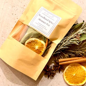 May include: A tan paper pouch with a clear window containing dried orange slices, bay leaves, and cinnamon sticks. The label reads Woodland Spice Simmer Pot, Grown-Harvested-Dried in Somerset. Beside the pouch are loose rosemary, cinnamon, and orange.