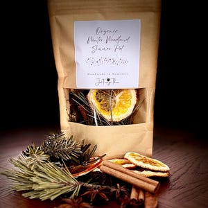 May include: A brown paper bag with a clear window showing dried orange slices, pine needles, and other botanicals. The bag is labelled "Organic Winter Woodland Simmer Pot" and "Handmade in Somerset". Cinnamon sticks and star anise are arranged around the bag.