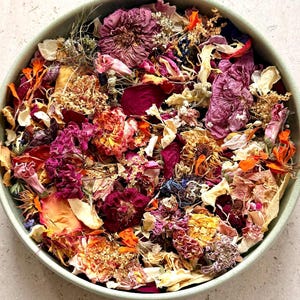 May include: A round, light green ceramic bowl filled with a dense mixture of dried flower petals and botanical elements. The assortment includes deep magenta, bright orange, soft yellow, and cream-coloured dried blossoms and foliage.