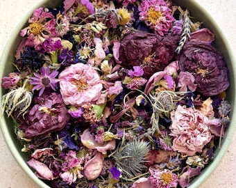 May include: A round, light green bowl filled with a dense mixture of dried flowers and botanicals in shades of deep purple, magenta, soft pink, and lavender. The collection includes dried rose heads, small petals, and spiky thistle elements.