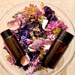 May include: A clear glass bowl filled with a mixture of dried flowers in shades of purple, pink, and white, featuring two small amber glass vials with black screw-top caps resting on top of the botanical arrangement.