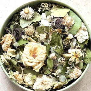 May include: A round, light green bowl filled with a collection of dried botanical elements, including cream-coloured roses, small white flowers, green eucalyptus leaves, and dark purple thistle heads, arranged in a dense, layered composition.