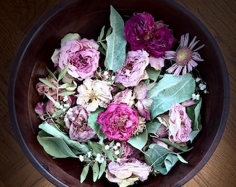 May include: A wooden bowl filled with dried flowers and greenery. The arrangement includes pink and burgundy roses, green leaves, and small white flowers. The bowl is dark brown and sits on a wooden surface.