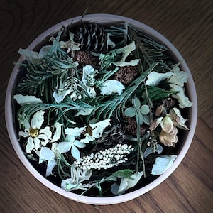 May include: A white bowl filled with a mixture of dried flowers, pine needles, and pine cones. The bowl is on a wooden surface.
