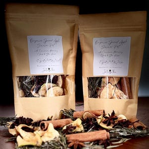 May include: Two brown paper pouches containing Organic Spiced Apple Simmer Pot with Cinnamon, Rosemary, Cloves & Star Anise. The labels state Handmade in Somerset and Just Forage Then. Dried apple slices, cinnamon sticks, and star anise are visible.