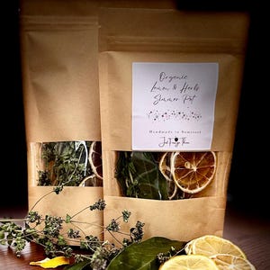 May include: Two brown paper bags with clear windows, filled with dried herbs and lemon slices. The bags have labels that read "Organic Lemon & Herbs Summer Pot". Fresh lemons, herbs, and peppercorns are arranged in front.