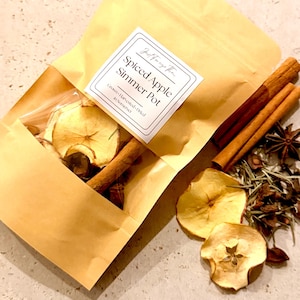 May include: A tan paper pouch labelled Spiced Apple Simmer Pot, Grown-Harvested-Dried in Somerset, containing dried apple slices and cinnamon sticks. Loose dried apple slices, cinnamon sticks, and star anise are arranged next to the pouch.