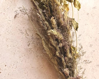 May include: A dried floral bouquet featuring a mix of golden-brown grasses, wheat stalks, and delicate dried leaves. The stems are gathered together and secured at the base with a simple, thin brown twine tied into a small, neat bow.