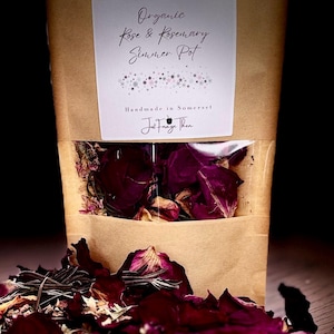 May include: A brown paper bag containing dried rose petals and rosemary, with a clear window showing the contents. The label reads "Organic Rose & Rosemary Summer Pot" and "Handmade in Somerset". Loose petals and herbs are scattered in front.