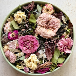 May include: A round, light green bowl filled with a collection of dried flowers and foliage. The arrangement features various shades of pink, cream, and white roses, interspersed with dark green and brown dried leaves and small textured botanical sprigs.